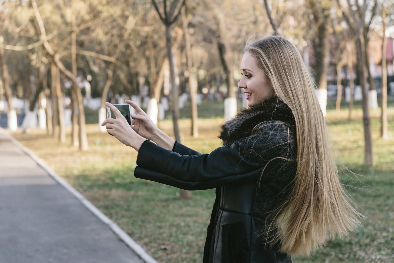 公園でスマートフォンを両手で構えて撮影する金髪ロングヘアの女性［モデル：モデルファクトリー］