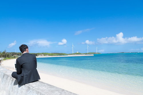 「バカンス中に何で修正依頼がくるんだよ！」と背中で愚痴るスーツ姿の男性