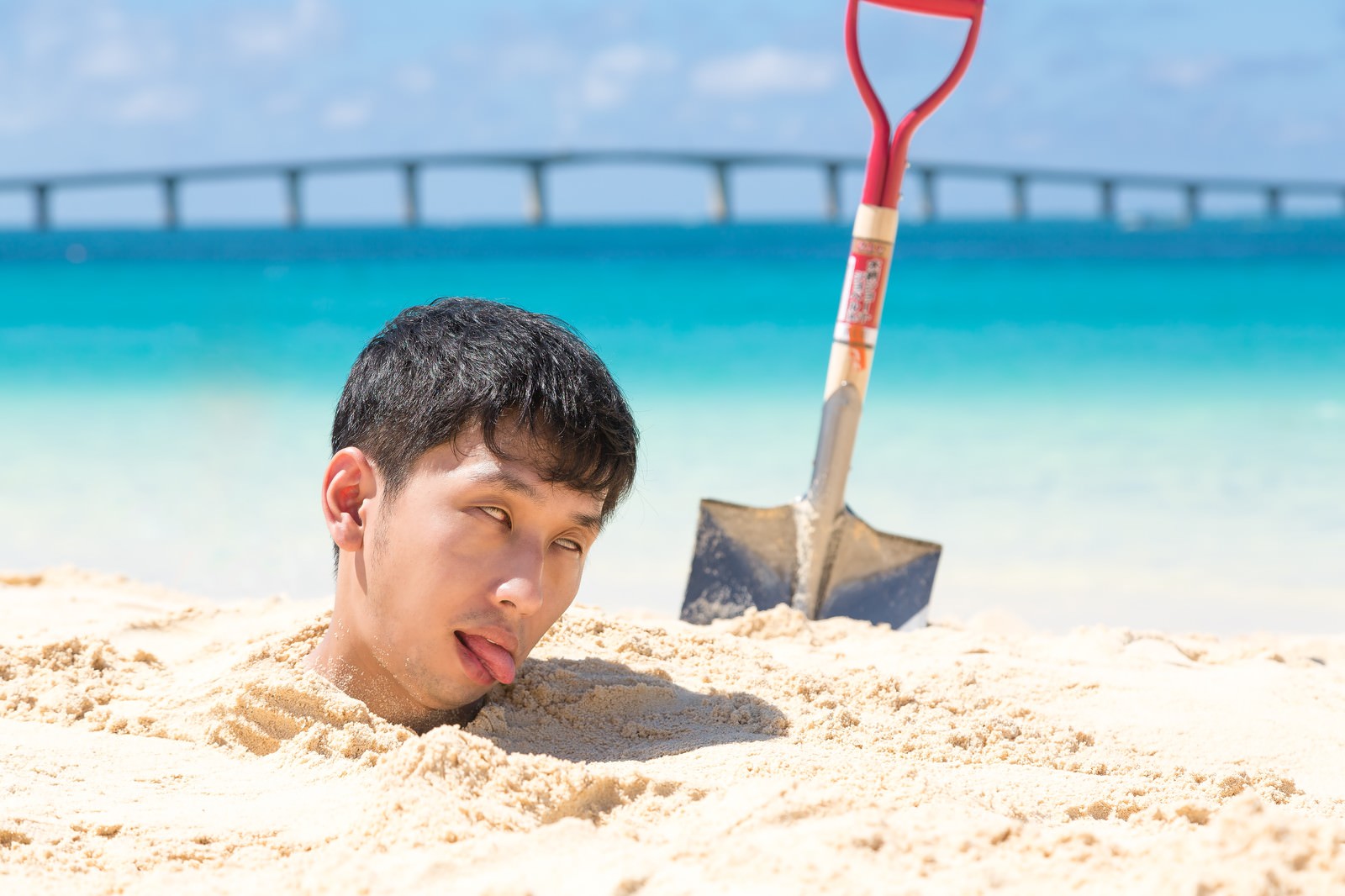 砂浜に首まで埋まりだるそうな表情の男性［モデル：大川竜弥］