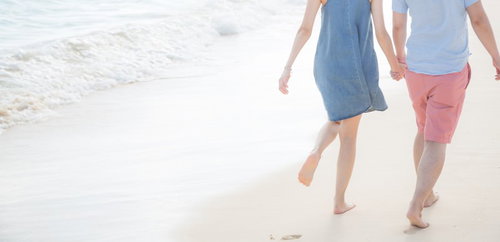 波打ち際の砂浜を手をつないで歩くカップルの後ろ姿
