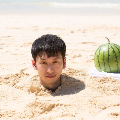 砂浜に首まで埋まった男性とスイカが並ぶ海辺のコミカル写真の写真