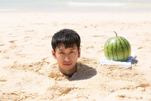 砂浜に首まで埋まった男性とスイカが並ぶ海辺のコミカル写真