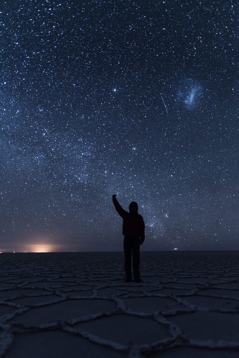 ウユニ塩湖の広大な塩湖に星が満天に輝き、一人の観光客が腕を上げて立っている夜間の風景