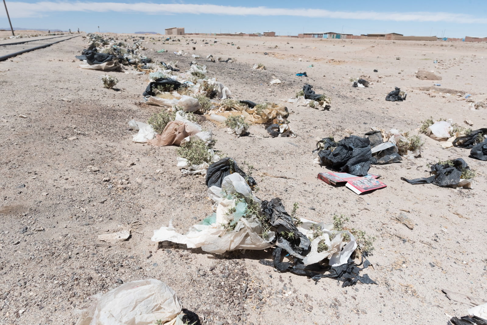 砂漠の茶色い地面に黒いビニール袋、白いプラスチック片、段ボール、布地が散乱している大量のゴミ