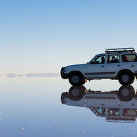 ウユニ塩湖の鏡面反射に映る白い4WD車両と乗員、アンデス山脈を背景にした光景の写真