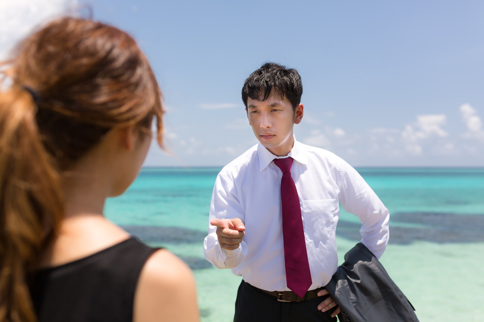 海辺で女性に人差し指を向ける白シャツの男性［モデル：大川竜弥］