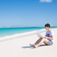 海で楽しそうに泳ぐ同僚を見ながら、ひざの上にMacBook Airを乗せて改修案件をするエンジニアの写真