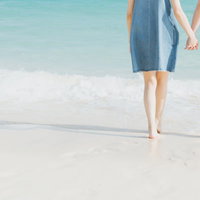 青い海の波打ち際で手をつなぐデニムワンピースのカップルの写真