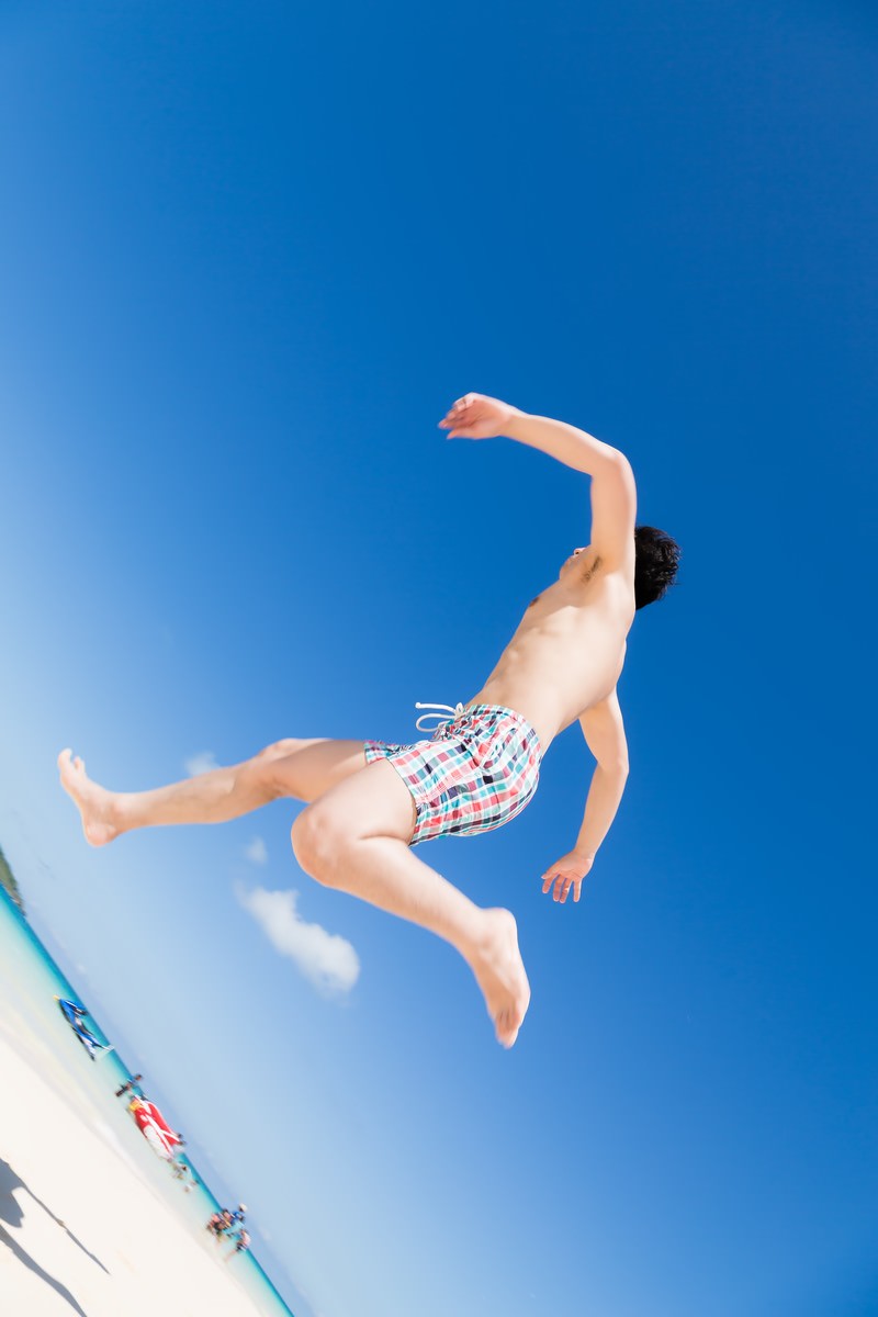 青空の下で飛び込み台からジャンプする海水パンツの男性［モデル：大川竜弥］
