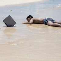 波打ち際に腹ばいで横たわる水着の漂流ノマド風男性の写真