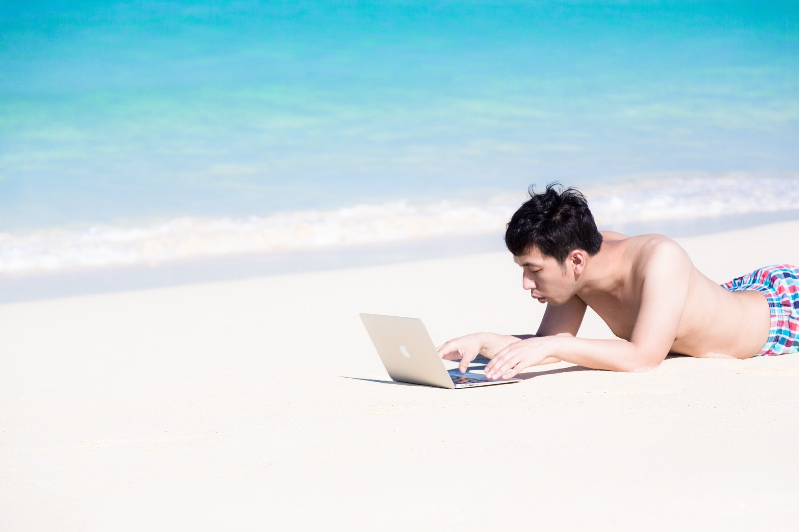 前浜ビーチの白い砂浜でうつ伏せにノートPCを操作する男性［モデル：大川竜弥］
