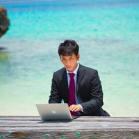 透明な海が見える木製桟橋に座ってノートPCを操作するスーツの男性の写真