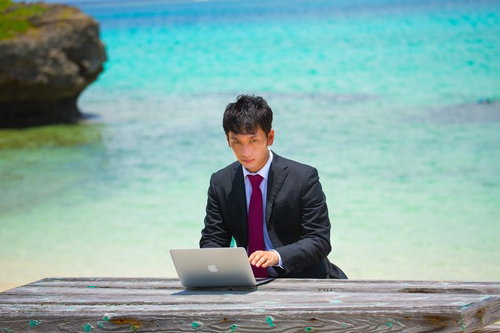 透明な海が見える木製桟橋に座ってノートPCを操作するスーツの男性