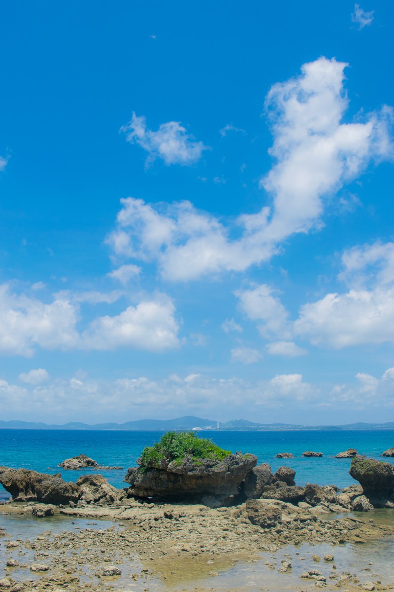 青い海に岩礁が点在し、白い雲が浮かぶ青空が広がる夏の海岸風景