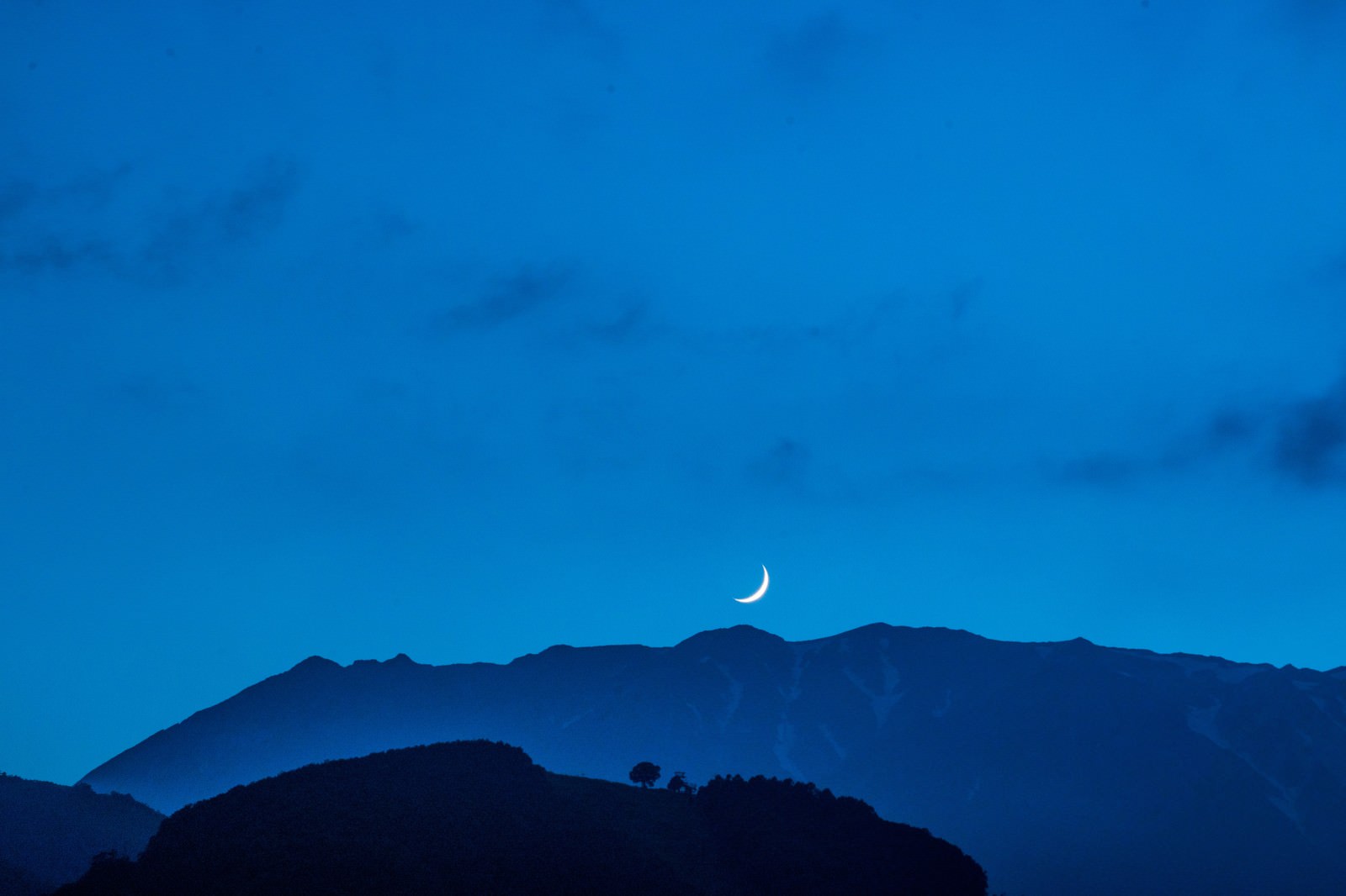 青い夜空に浮かぶ三日月と重なり合う山のシルエット