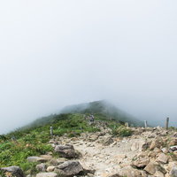 視界不良の雲の中へと進む登山者の後ろ姿と岩場の登山道の写真