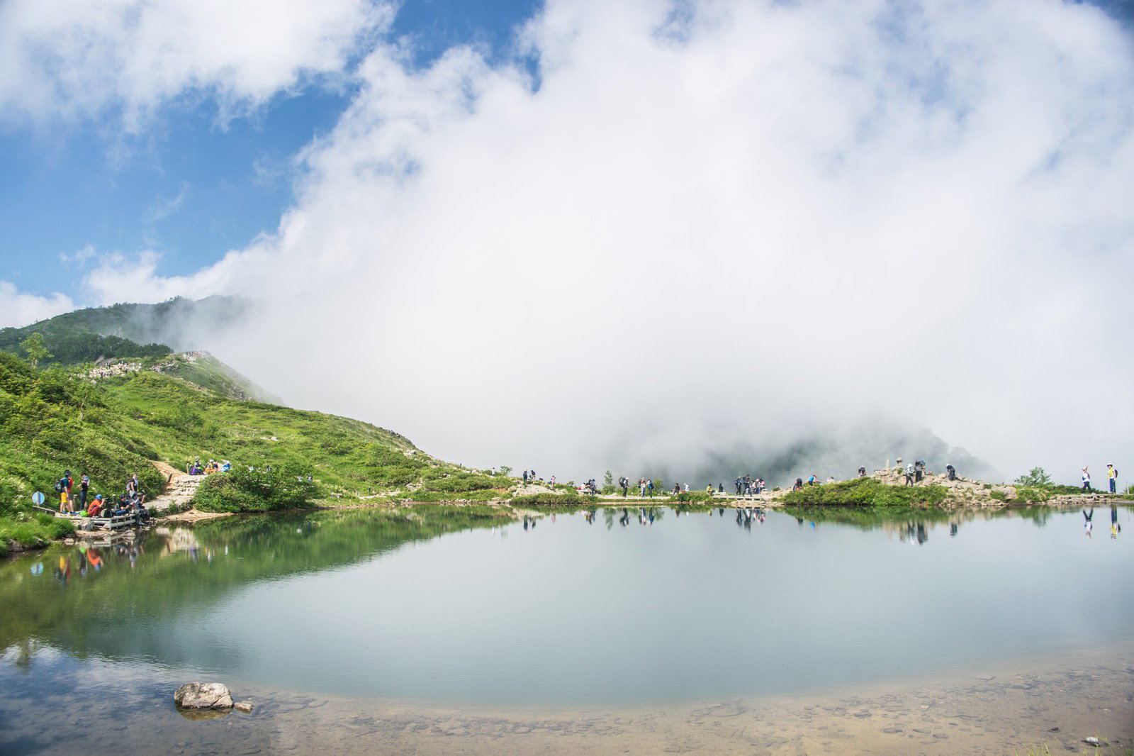 緑の山々に囲まれた湖で休憩する登山者たちと青空が映る湖面