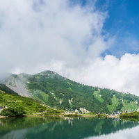 沸き立つ雲と湖面に映る山々を眺める登山者の写真