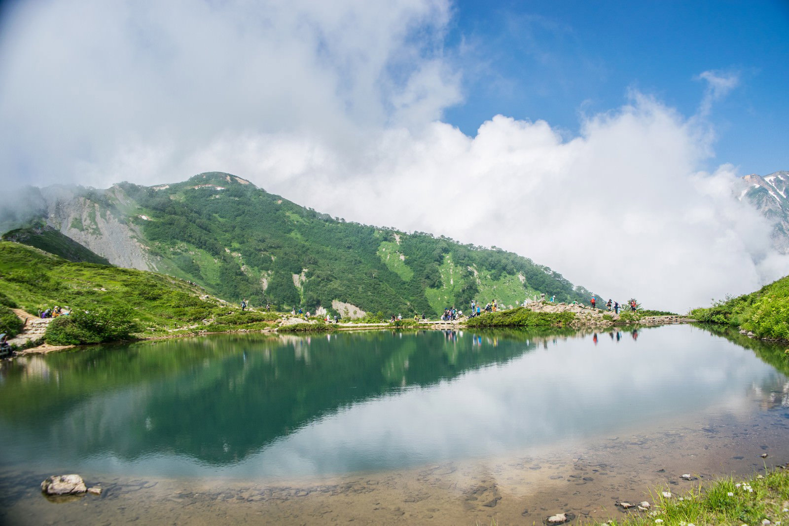 湖畔で休憩する登山者と湖面に映る緑の山々