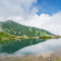 湖畔で休む登山者と水鏡に映る緑の山々、青空と雲の写真