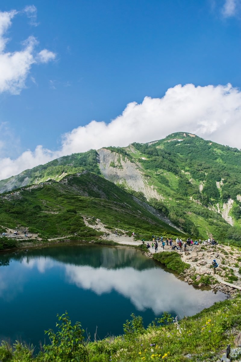 青空と雲が広がる山間の湖畔で、緑の山々を背景に登山者が佇む風景