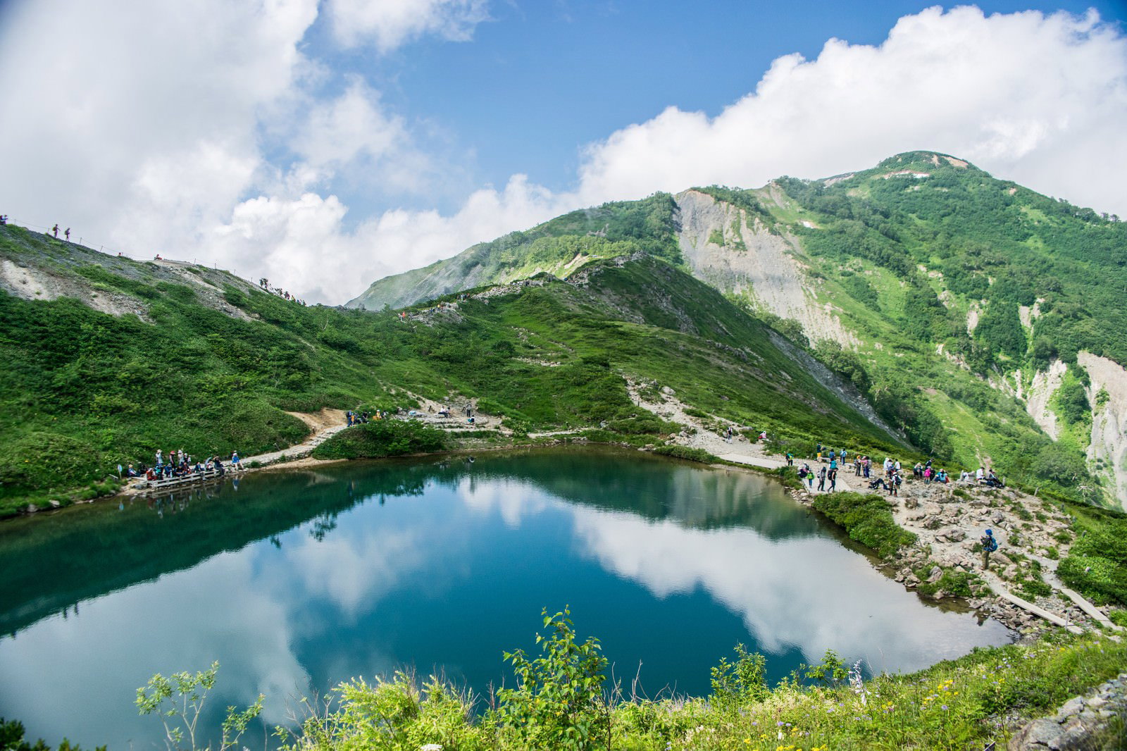 静かな湖面に青空と雲が映り込む水鏡と、緑の山々を背景にした登山者