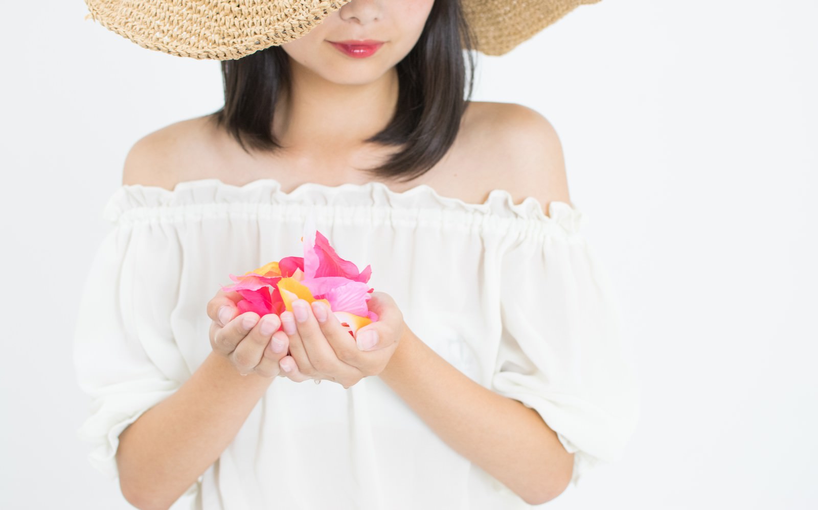 麦わら帽子をかぶった女性が白いオフショルダーの服を着てピンクの花を両手で持っている