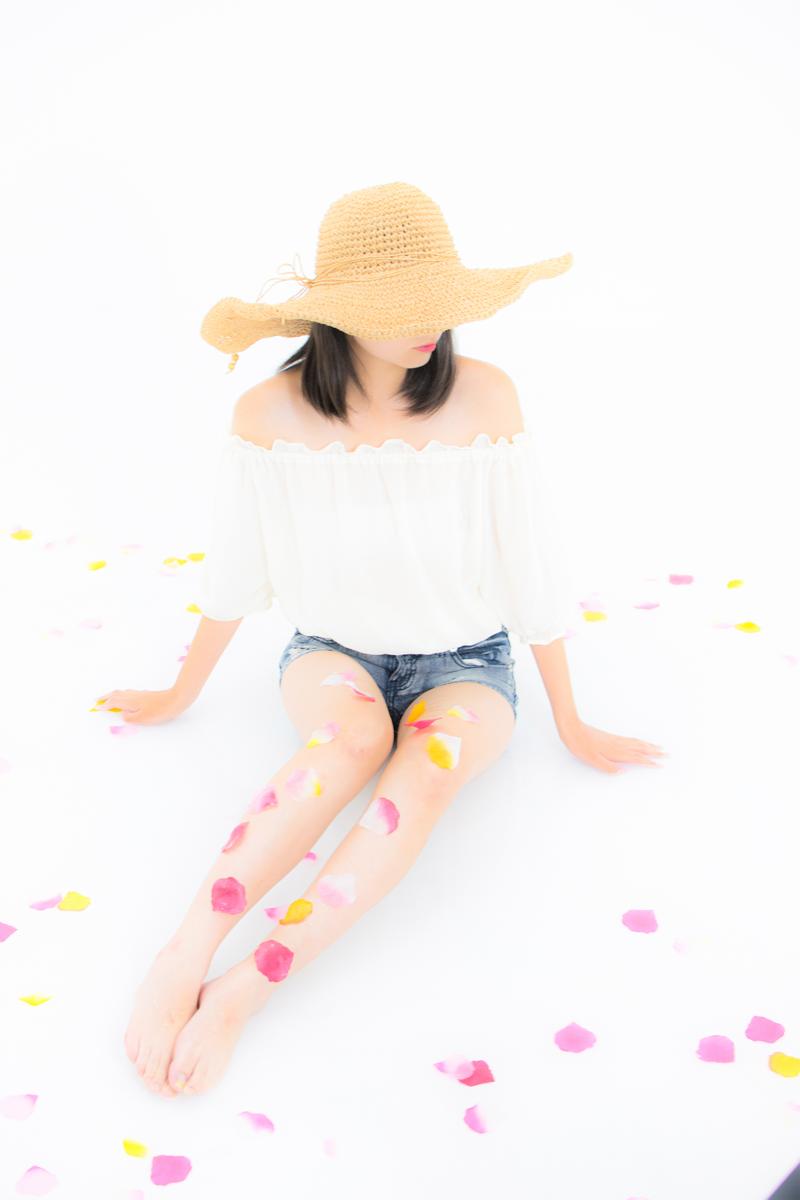 麦わら帽子をかぶった女性が白いオフショルダートップスとデニムショーツで座っている
