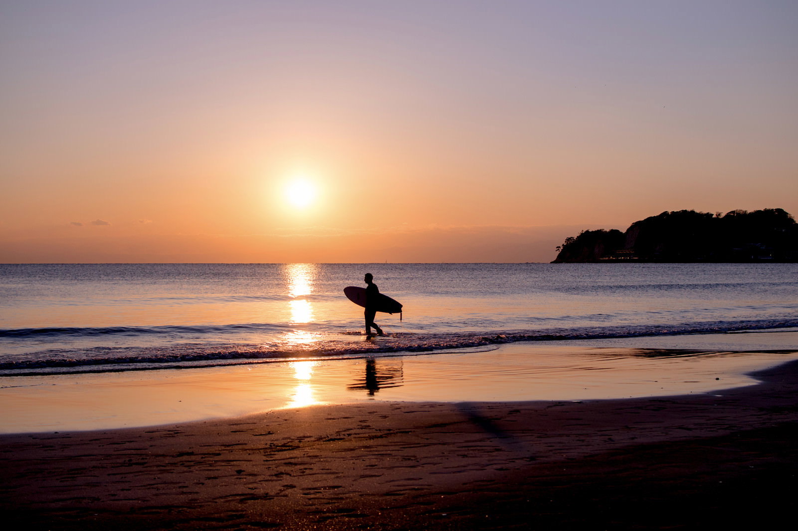 夕日を背景にサーフボードを持って波打ち際に立つサーファーのシルエット