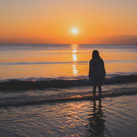 夕焼けに向かって浜辺に立ちすくむ女性のシルエットの写真