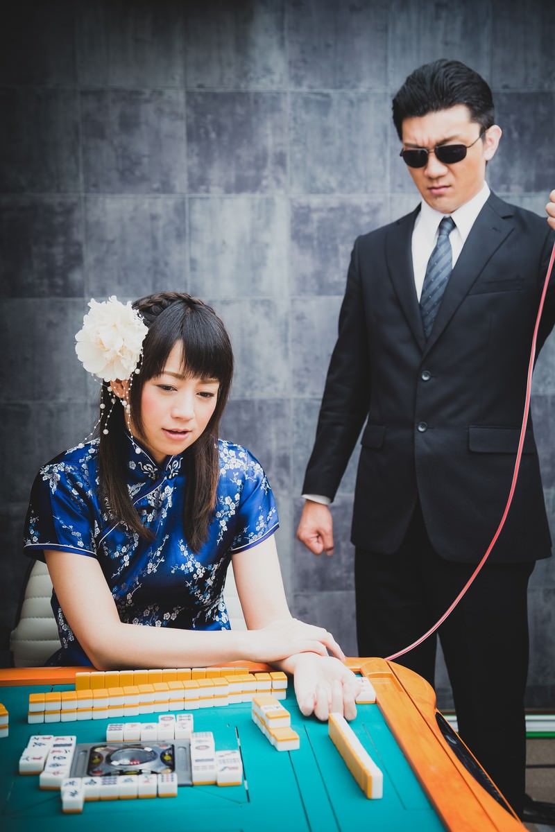 麻雀卓で点滴を受けながら牌を打つチャイナ服の女性と、サングラスの黒スーツ男性［モデル：米崎奈棋 OZPA］