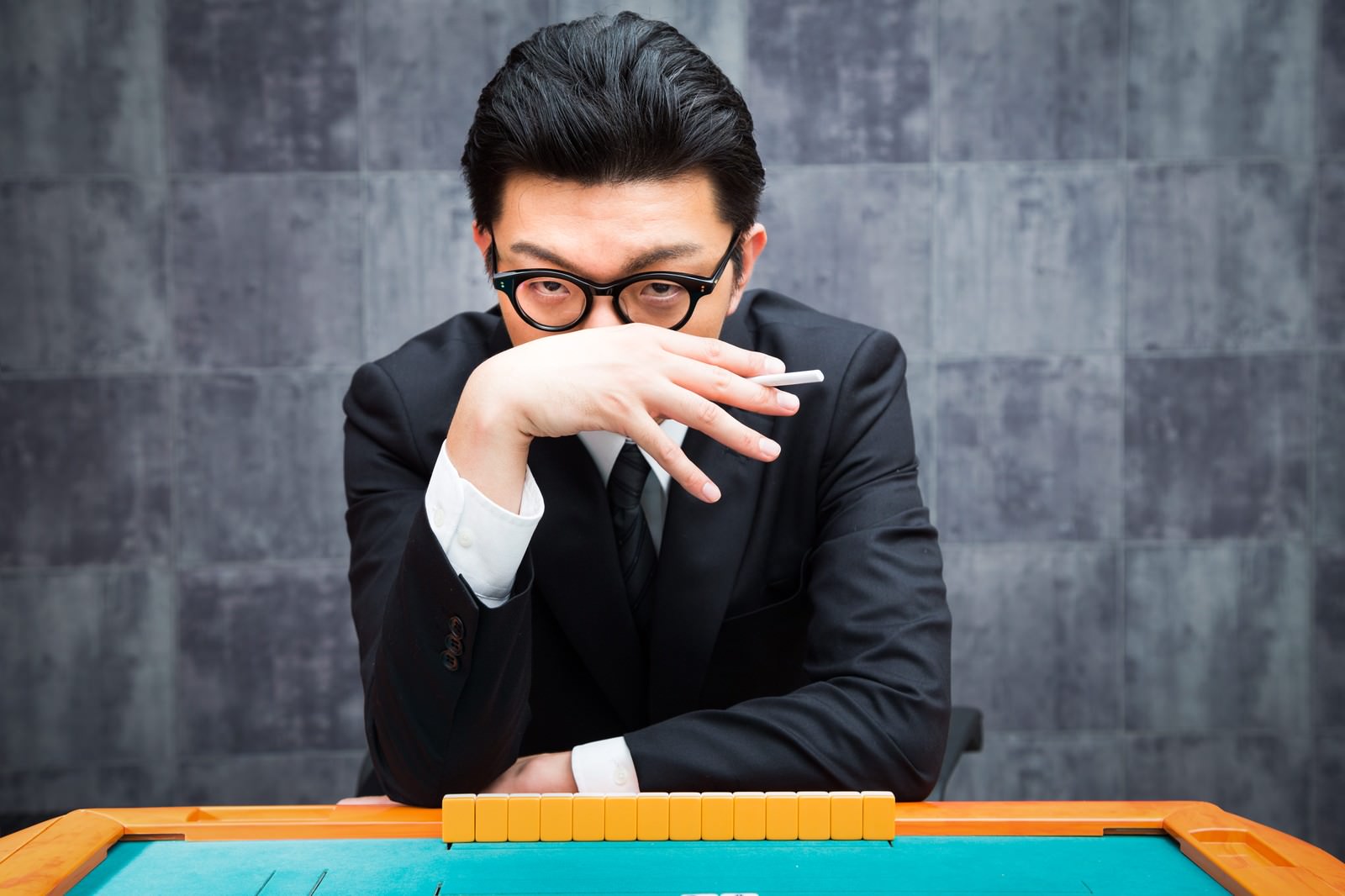 黒いスーツの男性が麻雀卓でリーチを仕掛け、真剣な表情で眼光を放つ［モデル：OZPA］