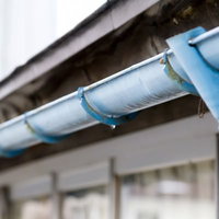 曇り空の下の民家の屋根に設置された青い雨どいと排水パイプの写真