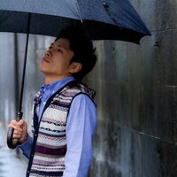 雨の日にコンクリート壁の前で紺色の傘を差す青年の写真