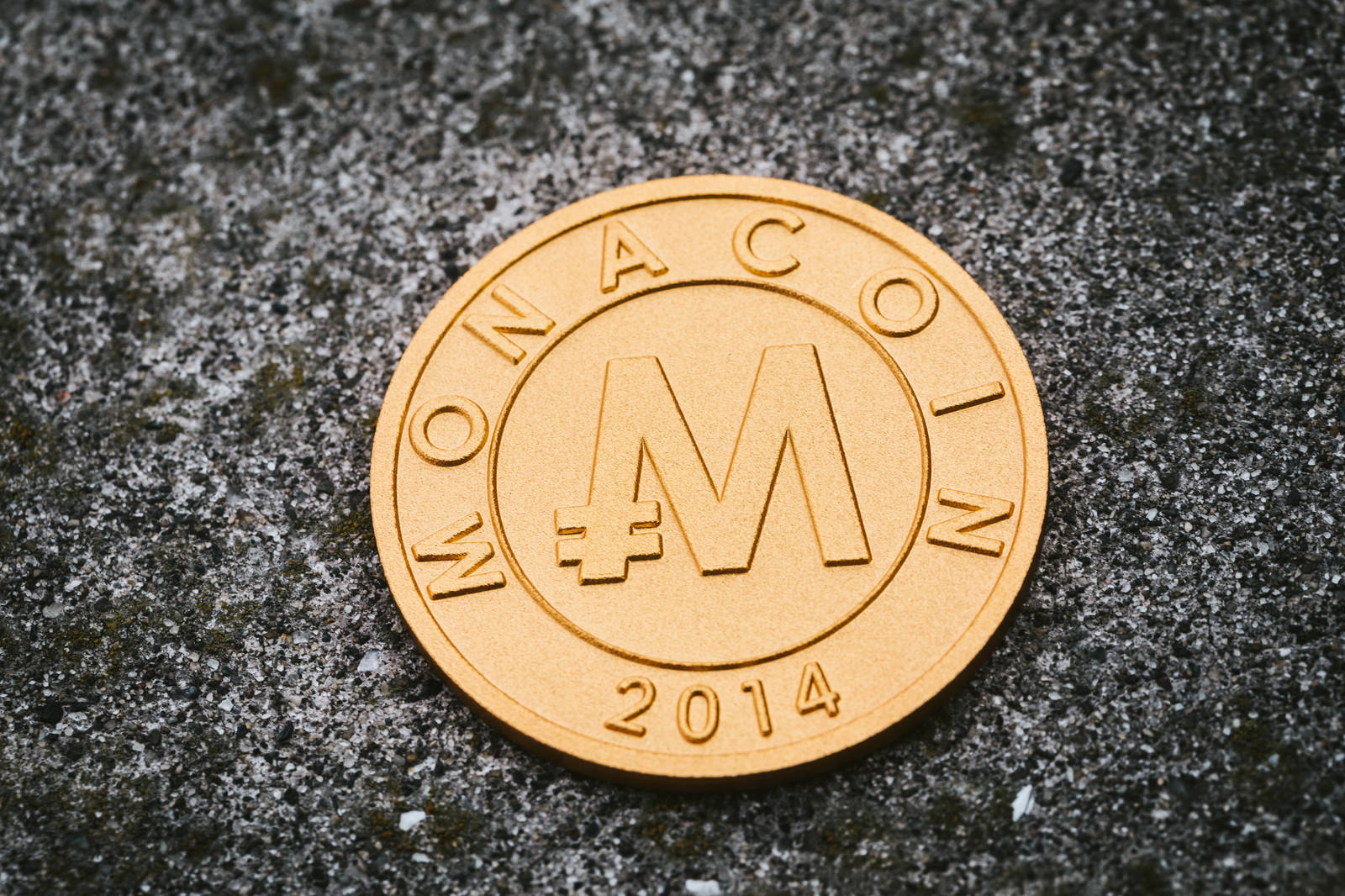 黒い石の背景に置かれた金色のモナコイン、表面にM刻印と2014の文字が見える