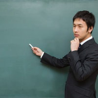 黒板に向かってチョークを当てたまま考え込む先生の後ろ姿の写真