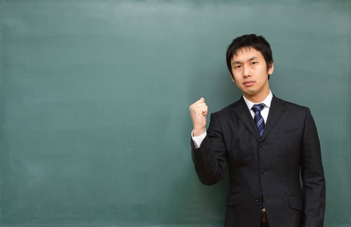 黒板の前で拳を握って「負けるな学生！」と応援する塾講師