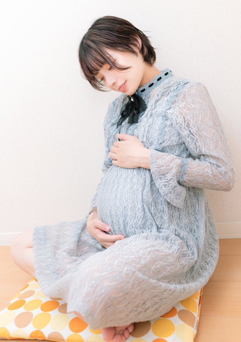 グレーのパジャマを着た妊婦が座ってお腹に手を当てている［モデル：高尾実生］