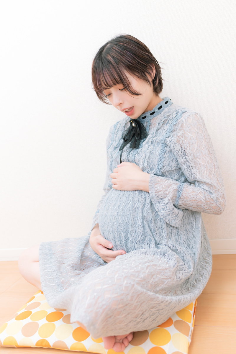 グレーのパジャマを着た妊婦が座ってお腹に手を当てている［モデル：高尾実生］