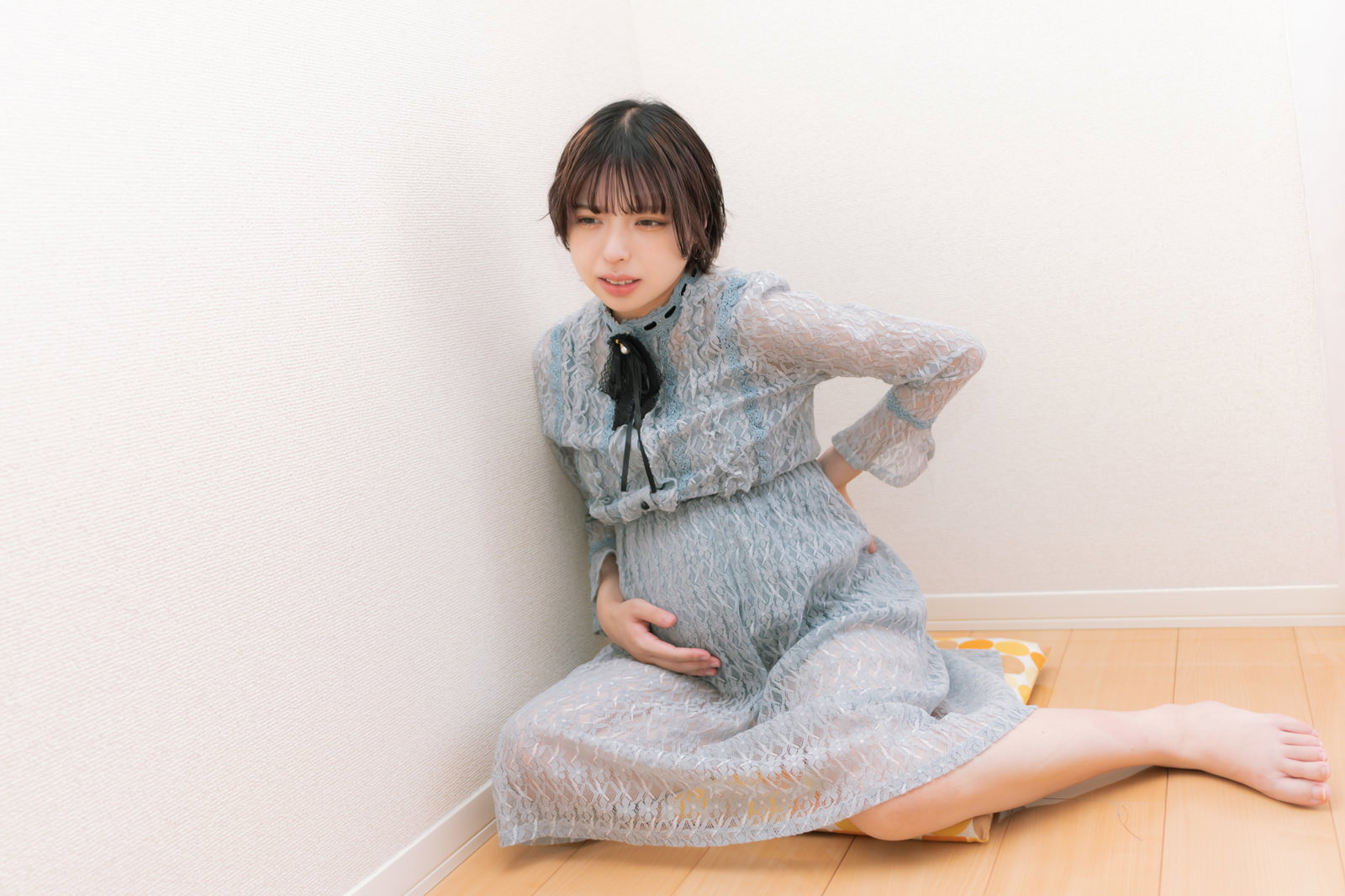 グレーのパジャマを着た妊婦が床に座り腰に手を当てて痛みを感じている［モデル：高尾実生］