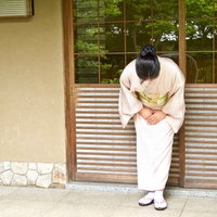 日本家屋の玄関前でお辞儀をする着物姿の女将さんの後ろ姿の写真
