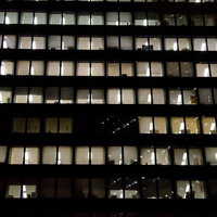 夜のオフィスビルの窓から漏れる明かりが並ぶ眠らないオフィスの写真