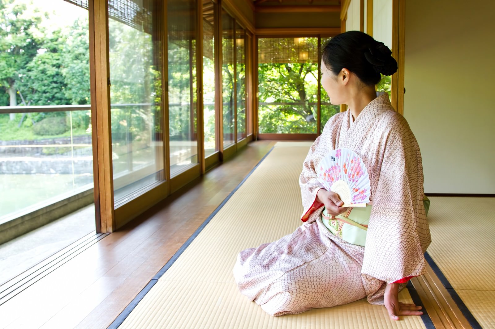 緑が見える廊下で扇子を持ち座る着物の女性［モデル：きせんひろみ］
