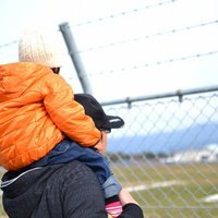 フェンスの前で子供を肩車する親子の後ろ姿の写真