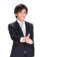 水色の壁の前で笑顔で手を差し出し握手を求めるスーツの男性の写真