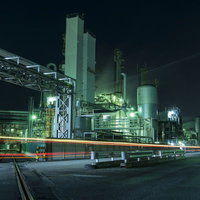 夜間に照明が点灯した工場の煙突と配管の写真