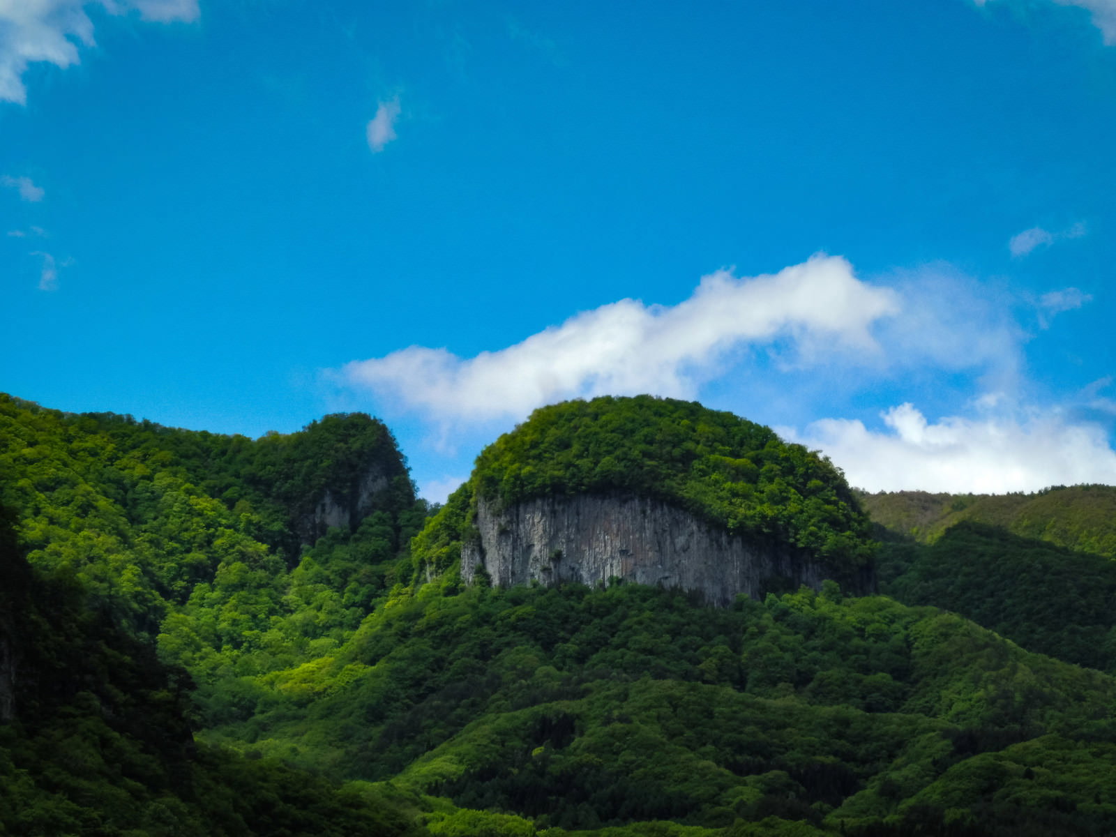 青空と白い雲をバックに緑の山々に囲まれてそびえ立つ絶壁の丸岩