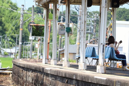 ローカル線の駅ホームで待つ爽夏、青いベンチに座る女性