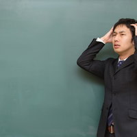 黒板の前で両手で頭を抱えて悩み込む塾の講師の写真