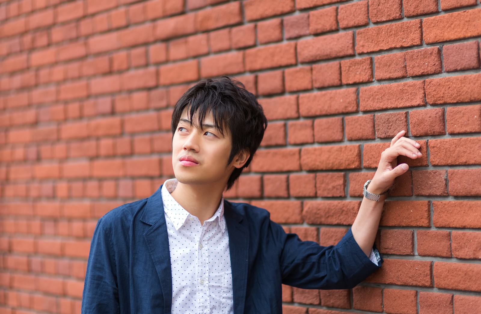 レンガ壁に寄りかかるカジュアルな青年［モデル：Tsuyoshi.］